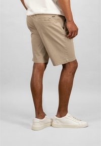 Beige puuvillashortsit suoran leikkauksen omaavina, joissa on huomaamaton vetoketjullinen tasku ja joita on yhdistetty valkoisiin lenkkareihin. Sileä pinta ja rento muotoilu.