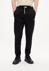 Man in zwarte tapered sweatpants met elastische tailleband en beige trekkoord, gecombineerd met zwarte sneakers, staand met zijn handen in zijn zakken.
