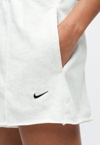 Άτομο που φοράει λευκά σορτς Nike, με το χέρι τοποθετημένο στην δεξιά τσέπη, δείχνοντας το μαύρο λογότυπο  της Nike κοντά στο τελείωμα.