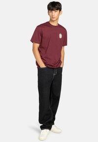 Camiseta de algodón burdeos con cuello redondo y un detalle de logotipo circular en el lado izquierdo del pecho, combinada con jeans negros holgados y zapatillas blancas.