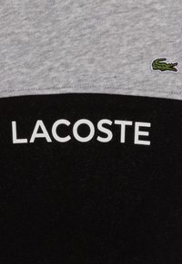 Graues und schwarzes Baumwollhemd mit einem kleinen grünen Krokodil-Logo und dem Aufdruck "LACOSTE" in Weiß auf dem schwarzen Teil.