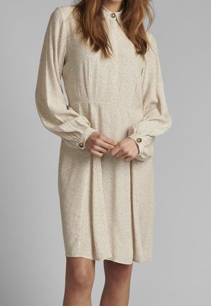 Kvinde iført beige langærmet kjole med lille prikket mønster, knapper ved manchetterne og en høj halsudskæring med en dekorativ knap.