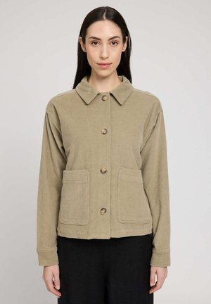 Woman with long dark hair wears beige button-up corduroy jacket with large front pockets and black pants, standing against plain background.