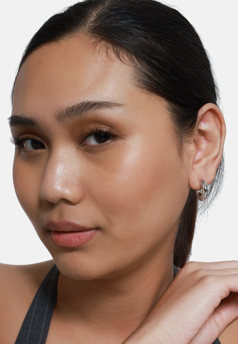 Primer plano de una persona con tono de piel medio, que lleva pendientes de aro plateados. El cabello es oscuro y liso, con un cutis natural y suave.