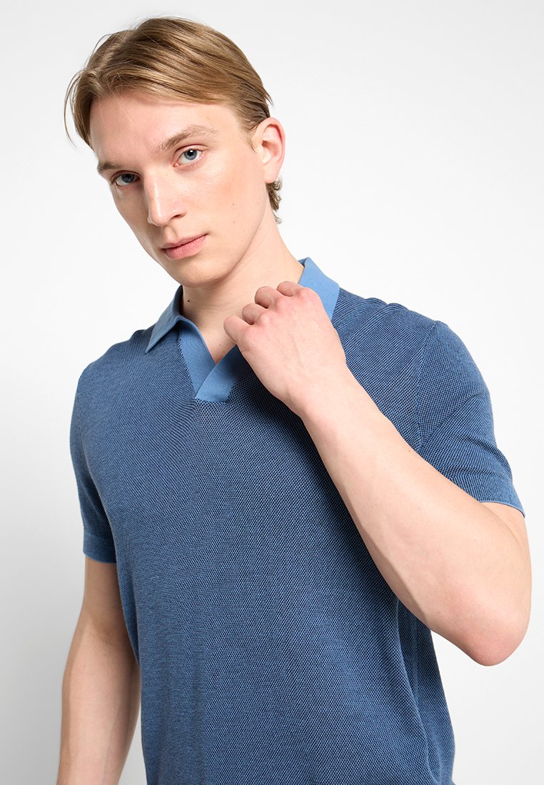 Blaues gestricktes Poloshirt mit einem kontrastierenden hellblauen Kragen, kurzen Ärmeln und einem strukturierten Muster. Modell, das den Kragen anpasst, vor einem neutralen Hintergrund.