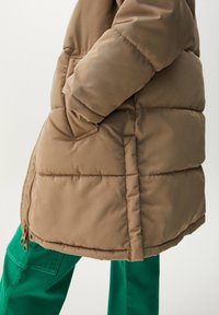 Veste matelassée marron avec texture matelassée, dotée d'une poche latérale et d'un détail zippé. La veste est associée à un pantalon vert.