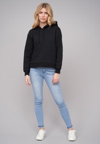 Felpa nera con cappuccio e cordini, polsini e orlo a costine, abbinata a jeans skinny azzurri e sneakers bianche, contro uno sfondo grigio.