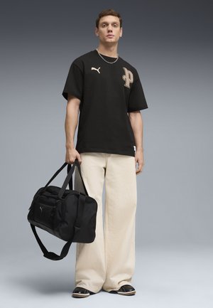 Junger Mann trägt ein schwarzes Puma-T-Shirt und beige weite Hosen, hält eine schwarze Puma-Reisetasche vor einem grauen Hintergrund.