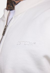 Weiße Zip-Jacke aus strukturiertem Stoff, mit einem geprägten "ESTEEM"-Logo auf der Brust und einem silbernen Reißverschluss.