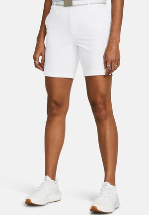 Persoon draagt witte shorts tot halverwege de dij, een witte riem, witte sneakers en ringen aan de vingers tegen een eenvoudige lichte achtergrond.