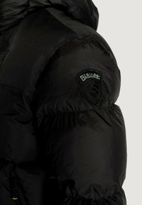 Giubbotto invernale nero con texture trapuntata, motivo cucito e una grande toppa con logo sulla spalla. Tessuto resistente con finitura lucida.