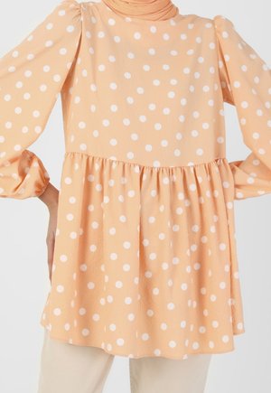 Lyserød langærmet bluse med hvide polkaprikker, sammenrynket talje og puffede ærmer båret med lyse bukser.