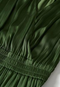 Tissu plissé vert avec une texture lisse, doté d'un élastique plié à la taille pour un design ajusté. La lumière se reflète subtilement sur la surface.