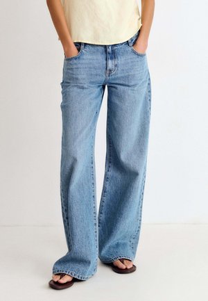 Person trägt hellblaue Jeans mit weitem Bein, cremefarbenes Hemd, das in die Hose gesteckt ist, und braune Sandalen, steht mit den Händen in den Taschen vor einem einfarbigen Hintergrund.