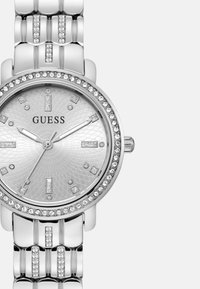 Silberne Guess-Uhr mit strukturiertem Zifferblatt, Diamantakzenten und mit Kristallen besetzter Lünette. Sie verfügt über ein armbandes Design aus verknüpften Metallgliedern.