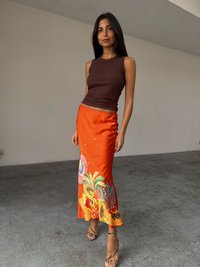 En kvinne står i et innendørs rom iført en tettsittende brun tanktopp og et livlig oransje skjørt med blomsterdesign, sammen med strappy sandaler.