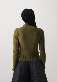 Oliven grønn ribbet langermet topp med bølgekant og tettsittende design, brukt med et mørkt quiltskjørt.