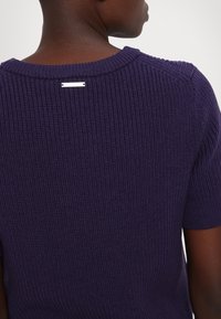 Fialový žebrovaný svetr s krátkými rukávy, kulatým výstřihem a malou kovovou značkou na zadní straně.