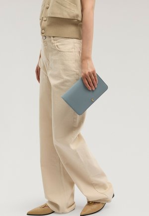 Persona con pantalones beige de pierna ancha, zapatos puntiagudos color tan y sosteniendo un bolso de mano azul con textura y un emblema dorado a un lado.