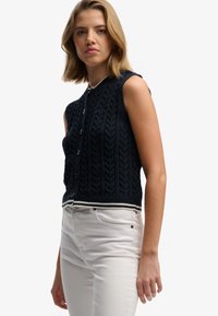 Chaleco de punto sin mangas azul marino con patrón de trenzas, cierre frontal con botones y ribete blanco contrastante en el dobladillo, combinado con jeans de color claro.