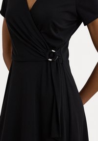 Zwarte wikkeljurk met korte mouwen, voorzien van een gedraaide knoopdetail in de taille en een zilveren ringaccent. Glad stof met een lichte rek.