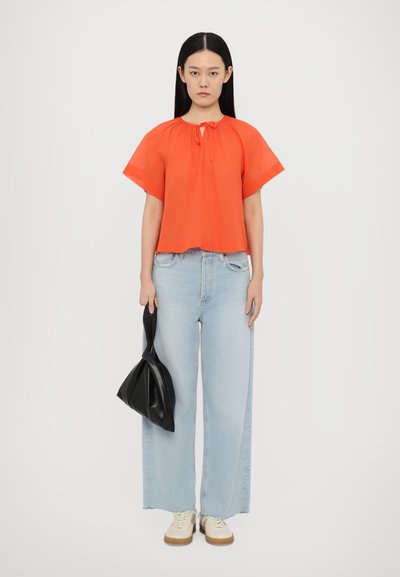 Orange Bluse mit kurzen Ärmeln, gesammeltem Halsausschnitt und Binder, kombiniert mit hellblauen weiten Jeans und einer schwarzen Handtasche.