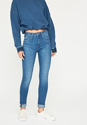 Frau, die blaue, umgeschlagene Skinny-Jeans, einen lockeren blauen Sweatshirt und weiße Sneaker trägt und vor einem einfarbigen Hintergrund steht.