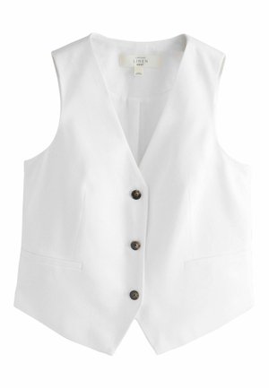 Femme en gilet blanc sans manches et pantalon large, debout au soleil contre un mur texturé, tenant une pochette noire à pois dans la main gauche.