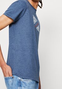 Persona vestida con una camiseta gráfica azul y jeans de color azul claro, con una mano en el bolsillo, frente a un fondo claro y simple.