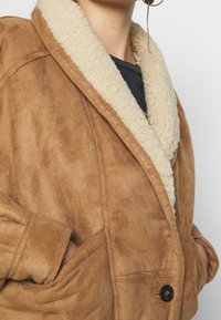 Brun faux suède jacka med en krämfärgad faux pälskrage, sidofickor och knappstängning. Har en avslappnad passform och mjuk textur.