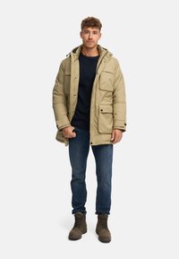 Beige isolert parka med hette, flere frontlommer, svart tilbehør, båret over en marineblå genser, kombinert med blå jeans og brune støvler.