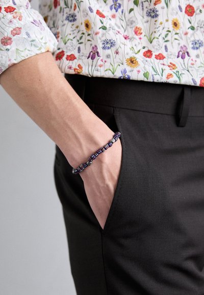 Bracelet en perles avec des composants bleu marine, rouge et argent, porté au poignet. La main est glissée dans la poche d'un pantalon noir.