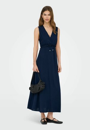 Kobieta w granatowej sukience maxi bez rękawów z paskiem w talii, trzyma czarną torebkę i nosi czarne buty typu Mary Jane, stoi na białym tle.