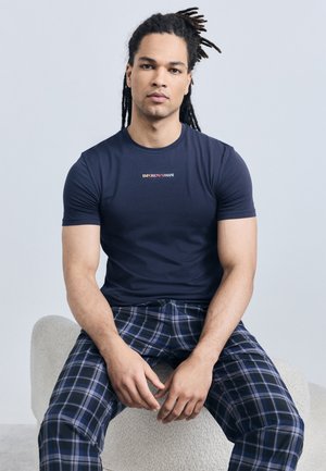 Άνδρας με πλεξούδες που κάθεται σε καμπυλωτό, υφασμάτινο σκαμπό, φοράει μπλε μπλουζάκι Emporio Armani και μπλε παντελόνι καρό.