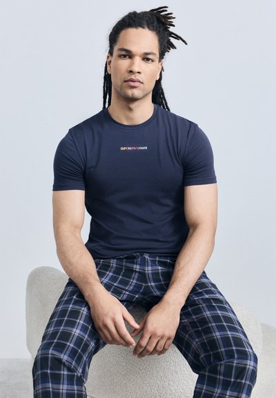 Homme avec des dreadlocks assis sur un tabouret incurvé et texturé, portant un t-shirt Emporio Armani bleu marine et un pantalon à carreaux bleus.