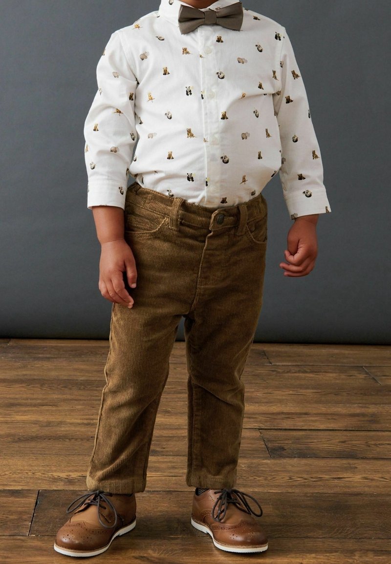 Jeune enfant portant une chemise blanche à imprimé animal, un pantalon en velours côtelé marron, un nœud papillon assorti et des chaussures habillées marron sur un sol en bois.