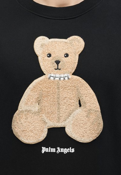 Εφαρμογή αρκουδάκι μπεζ με περιλαίμιο από ασημένιο παραμάνα σε μαύρο ύφασμα, κείμενο "palm angels" κάτω σε λευκή γοτθική γραμματοσειρά.