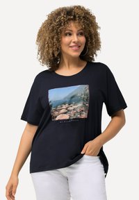 Námorní t-shirt s krátkým rukávem v námořnické modři, zdobený grafickým potiskem s přímořským motivem a třepeným okrajem. Hladká textura, volný střih, kombinovaný s bílými kalhotami.