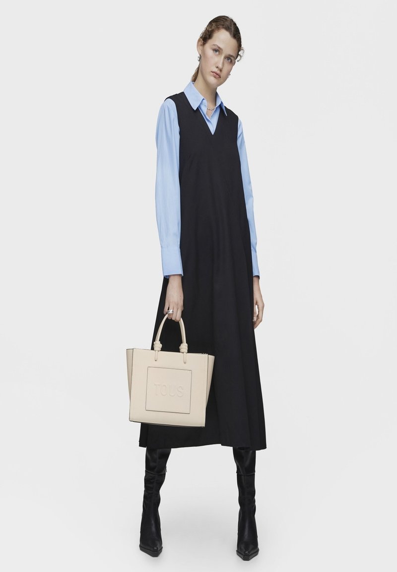 Czarna sukienka bez rękawów na niebieskiej koszuli z kołnierzykiem, w połączeniu z beżową skórzaną torebką z wytłoczonym logo. Czarnekozaki do kolan.