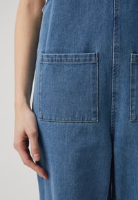 Denim salopete vsebujejo dva sprednja žepa, modro barvo in vidno šivanje. Tkanina ima mehko teksturo z rahlim sijajem.