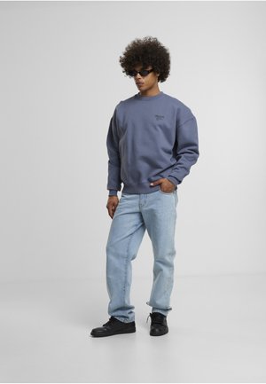 Man with curly hair wearing black sunglasses, blue sweatshirt, light blue jeans, and black sneakers standing against plain gray background.