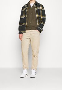 Codzienny strój składający się z czarno-żółtej koszuli flanelowej w kratę, oliwkowo-zielonej dzianinowej kamizelki, beżowych spodni oraz białych tenisówek z czarnymi detalami.