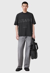 Un tânăr îmbrăcat într-un tricou oversize negru AllSaints, blugi gri cu croi dreapt, pantofi negri și ținând o geantă neagră cu mânere.