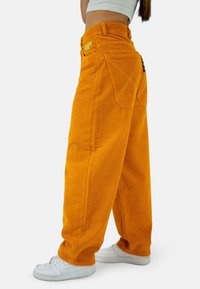 Oranssipintaiset corduroy-housut, joissa on leveät lahkeet, ommellut koristeet ja kaksi takataskua. Yhdistetty valkoisiin tennareihin.