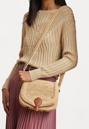 Vrouw draagt een beige kabelgebreide trui en een geplooide mauve rok, houdt een geweven strooien crossbodytas met leren sluiting vast.