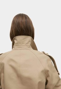 Beige trenchcoat met een hoge kraag, voorzien van een gestructureerd ontwerp en zichtbare stikdetails. De achteraanzicht toont een gladde stoftextuur.