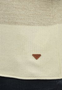 Pull en tricot épais beige et crème avec un motif texturé. Comprend une étiquette triangulaire en cuir marron à l'ourlet.