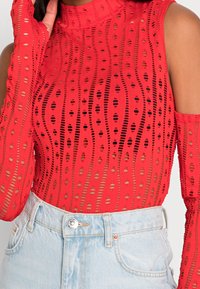 Red long-sleeve top with a high neck, featuring cut-out patterns in wavy lines. Worn with light denim jeans. Textured, breathable fabric.