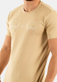 Camiseta de algodón beige de manga corta, con texto bordado en blanco "Project x Paris" en el pecho, textura suave y diseño ajustado.