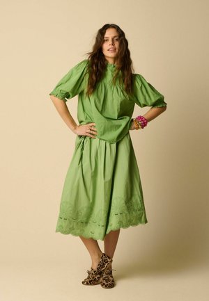 Femme debout, mains sur les hanches, portant une robe verte ample avec un ourlet festonné et des chaussures imprimé léopard, fond beige.